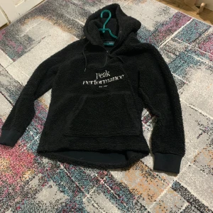 Svart Peak Performance hoodie i fleece - Mysig svart hoodie från Peak Performance med broderad logga på bröstet. Tröjan är tillverkad i fluffig fleece och har huva med snörning samt en stor magficka. Ribbstickade muddar vid ärmslut och något längre baktill för extra komfort.