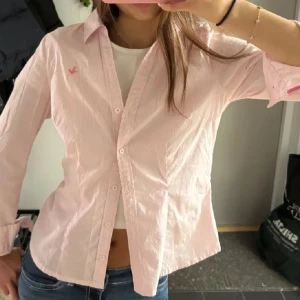 Rosa randig skjorta  - Snygg rosa skjorta från Broadway NYC Fashion i storlek S. Skjortan har tunna vita ränder, klassisk krage och knappar framtill. Detalj med rosa broderad logga på bröstet och rosa kant på insidan av kragen. Perfekt till jeans för en fräsch look.
