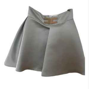 Blå kortkjol Elisabetta Franchi - Supersnygg grå kortkjol från Elisabetta Franchi med A-linjeformad siluett och exklusivt metallspänne framtill. Kjolen har en stilren design, dragkedja bak och är tillverkad i polyester med ett mjukt satinliknande foder. Perfekt för dig som gillar clean och modern look.