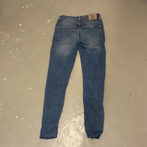 Blå jeans från Nudie Jeans - Säljer ett par klassiska blå jeans från Nudie Jeans med snygga kontrastsömmar och ikonisk broderad bakficka. Modellen är slim och skinny med normal midja och tillverkad i mjukt bomullsjeans. Perfekta för dig som gillar en clean och tidlös look.