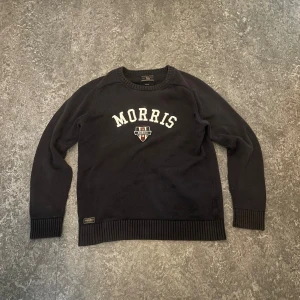 Svart stickad tröja från Morris - Svart stickad tröja från Morris med vit logotyp och emblem på bröstet. Tröjan har rund hals, ribbade muddar vid ärmslut och nederkant samt långärmad design. Perfekt för dig som gillar klassisk och sportig stil.