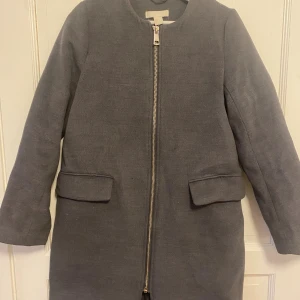 Grå rock med dragkedja från H&M - Snygg grå rock från H&M med rak passform och stilren design. Jackan har en hel dragkedja framtill, två stora fickor med lock och rundad halsringning. Perfekt för dig som gillar minimalistisk stil och vill ha en enkel men trendig jacka.