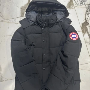 Canada Goose Wyndham - Svart dunjacka från Canada Goose med ikonisk röd och vit patch på ärmen. Jackan har huva, dragkedja och knappar framtill, samt quiltad design för extra värme. Perfekt för kalla dagar och har en clean, stilren look som funkar till streetwear. Pris kan diskuteras
