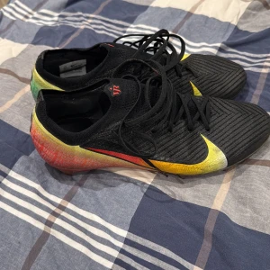 Svarta fotbollsskor (VINI JR) ELITE - Säljer ett par svarta Nike fotbollsskor med snörning och regnbågsfärgad häl i rött, gult och grönt. Skorna har en stickad ovandel med VJR-logga på plösen och textdetaljer på hälen. Snyggt mönstrad yta och platt sula för grym känsla på planen.
