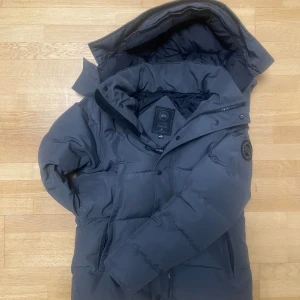 grå jacka Canada Goose - Säljer en mörkgrå dunjacka från Canada Goose med stor huva och snyggt svart patch på ärmen. Jackan har dragkedja och knappar framtill, två sidofickor och quiltad design. Perfekt för kalla dagar och riktigt stilren look. Den saknar en knapp som inte är visad i bilden