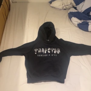 Svart hoodie från Trapstar - Svart hoodie från Trapstar med stor vit och grå logga på bröstet och texten 'IT'S A SECRET' under. Klassisk känguruficka framtill, huva med snörning och långa ärmar. Perfekt för dig som gillar streetwear och vill sticka ut med en snygg logga.