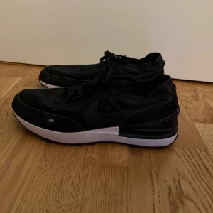 Svarta Nike sneakers med vit sula - Svarta Nike sneakers med klassisk låg siluett och vit platt sula. Skorna har snörning och är tillverkade i en kombination av syntet och skinn, med diskret Nike-logga på sidan. Perfekta för dig som gillar stilrena och sportiga skor.