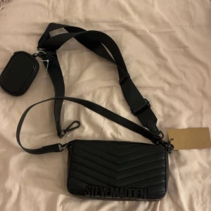 Svart quiltad axelväska Steve Madden - Snygg svart axelväska från Steve Madden med quiltad design och bred justerbar rem. Väskan har ett stort fack med magnetlås och en mindre avtagbar ficka på remmen. Perfekt för att bära dina essentials och ge din outfit en cool touch.