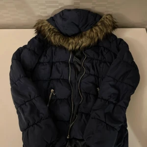 Mörkblå pufferjacka med fuskpäls - Säljer en mörkblå pufferjacka med huva och fuskpäls runt kragen. Jackan har quiltad design, två fickor med dragkedja och en rejäl dragkedja framtill. Perfekt för kalla dagar och har en riktigt mysig vibe.