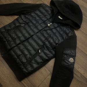 Moncler cardigan - Svart pufferjacka från Moncler med quiltad framsida, huva och dragkedja. Jackan har två fickor med dragkedja och en Moncler-logga på ärmen. Materialmix med polyester och syntet, perfekt för dig som gillar streetwear och snygga detaljer.