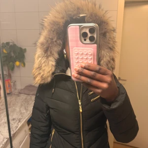 Svart pufferjacka med pälsluva - Säljer en svart pufferjacka från Hollies Stockholm med fluffig päls runt luvan och snygga guldfärgade dragkedjor. Jackan har flera fickor med dragkedja och en patch på ärmen. Perfekt för vinter och har en riktigt schysst look.