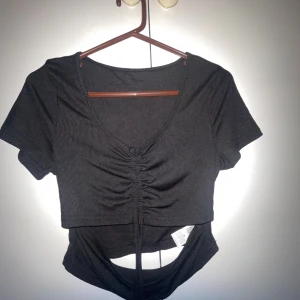 Svart croppad topp med snörning - Snygg svart croppad topp med ribbad struktur och v-ringning. Toppen har korta ärmar, snörning framtill och en cool cut-out detalj under bysten. Perfekt för dig som gillar trendiga och figurnära plagg med det lilla extra.