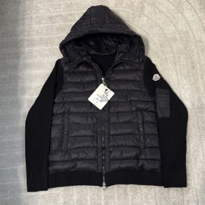 Svart Moncler pufferjacka med huva - Svart pufferjacka från Moncler i storlek M med huva och ribbstickade ärmar. Jackan har dragkedja framtill, två fickor med knappstängning och en extra ficka på ärmen med Moncler-logga. Insidan har ett coolt serietryck och märkestaggar. Perfekt för dig som gillar stil och funktion.