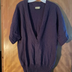 Lila stickad v-ringad tröja MyWear - Säljer en lila stickad tröja från MyWear med djup v-ringning och korta ärmar. Tröjan har bred ribbad mudd nedtill och runt halsen, vilket ger en snygg och avslappnad look. Perfekt för lager-på-lager-stil.