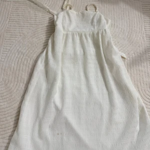 Vit kort sommarklänning med smala axelband - Säljer en vit kort klänning med smala axelband och lätt struktur i tyget. Klänningen har en luftig passform och är perfekt för varma dagar. Materialet känns mjukt och har ett diskret mönster som ger extra stilpoäng. Enkel och fräsch look!