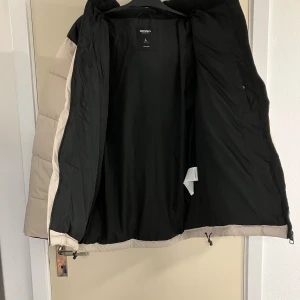 Beige och svart pufferjacka Icono - Säljer en beige och svart pufferjacka från Icono Couture med huva och dragkedja framtill. Jackan har en schysst tvåfärgad design, stora fickor med dragkedja och justerbar nederkant med snörning. Perfekt för kalla dagar när du vill hålla stilen.