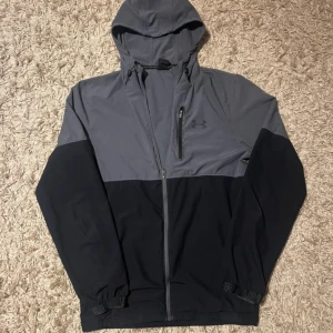 Grå och svart vindjacka Under Armour - Snygg vindjacka från Under Armour i grått och svart med huva och dragkedja framtill. Jackan har en bröstficka med dragkedja och elastiska muddar. Perfekt för dig som gillar sportig stil och vill ha något funktionellt och cleant.