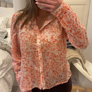 Blommig skjorta från Holly & Whyte - Säljer en söt blommig skjorta från Holly & Whyte by Lindex i storlek 38. Skjortan är vit med orange och röda blommor, har långa ärmar, knappar framtill och en krage med volangdetalj.
