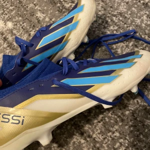 Adidas Messi blå fotbollsskor - Säljer ett par Adidas Messi fotbollsskor i blått med guld och ljusblå detaljer. Skorna har snörning, platt sula med dobbar och en strumpliknande krage för extra komfort. Tillverkade i syntetmaterial med coolt grafiskt mönster och Messi-logga på insidan.