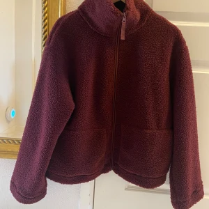 Vinröd teddyjacka med dragkedja från HM - Mjuk vinröd pilejacka med hög krage och två stora fickor framtill. Jackan har en rak, loose passform och stängs med dragkedja. Från H&M i storlek L.  Endast provad och är i nyskick. 