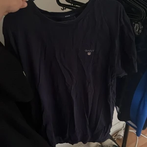 Mörkblå GANT t-shirt i bomull - Säljer en stilren mörkblå t-shirt från GANT i storlek M. T-shirten har klassisk rund hals, korta ärmar och en liten GANT-logga broderad på bröstet. Materialet är mjuk bomull och passformen är regular. Perfekt för dig som gillar enkla och snygga plagg.