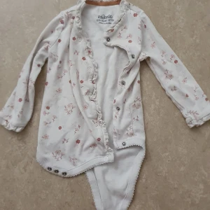Babybody - Supersöt vit babybody från So Cute i 100% ekologisk bomull. Långärmad med ljusrosa blommigt mönster, volangdetaljer längs knäppningen och ärmslut. Praktiska tryckknappar framtill och i grenen för enkel påklädning.
