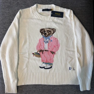 Vit stickad tröja Polo Bear Ralph Lauren - Vit stickad tröja från Polo Ralph Lauren med ikoniska Polo Bear-motiv i rosa och blått på framsidan. Rund halsringning och ribbade muddar vid ärmslut och nederkant. Tröjan är i bomull och har långärmad passform, perfekt för dig som gillar klassisk stil med en twist.