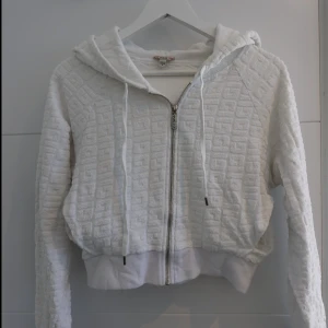 Vit hoodie från Guess med struktur - Säljer en vit hoodie från Guess med snyggt strukturerat mönster i tyget. Tröjan har dragkedja framtill, huva med snörning och breda muddar. Perfekt för dig som gillar en chill och trendig stil.
