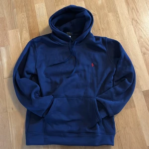Polo Ralph lauren hoodie - Säljer en marinblå hoodie från Polo Ralph Lauren med klassisk röd logga broderad på bröstet. Tröjan har huva med snörning, känguruficka och är tillverkad i mjuk bomull. Perfekt för dig som gillar stilren och rik stil. Hoodien är helt ny använd 1 gång hemma bara säljer för att den är för stor