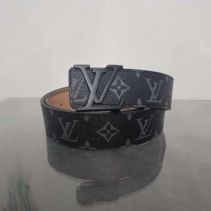 Svart Louis Vuitton bälte - Snyggt svart bälte från Louis Vuitton med klassiskt monogrammönster och en stor LV-logga som spänne. Bältet är gjort i skinn och har en stilren, mörk look som passar till jeans eller kostymbyxor. Perfekt för dig som vill ha en exklusiv accessoar.