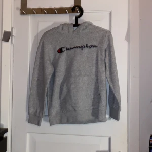 Grå hoodie från Champion - Grå hoodie från Champion med broderad logga i blått och rött framtill. Klassisk modell med huva och långa ärmar, ribbade muddar och mjukt bomullsmaterial. Perfekt för chill dagar eller när du vill ha en enkel och stilren look.