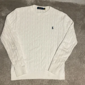 Vit kabelstickad tröja Polo Ralph Lauren - Vit kabelstickad tröja från Polo Ralph Lauren med klassisk rund halsringning och blå broderad logga på bröstet. Tröjan har långa ärmar och ribbade muddar för en snygg passform. Perfekt för dig som gillar stilren och tidlös design.