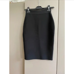 Svart pennkjol från F&P med dragkedja NY - Snygg svart pennkjol från F&P med markerad midja och synlig dragkedja baktill. Kjolen har en tight passform och är tillverkad i ett strukturerat syntetmaterial som ger en stilren look. Perfekt för dig som gillar minimalistisk och modern stil. Finns även i beige 