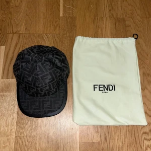 Svart Fendi keps med FF-mönster - Säljer en svart keps från Fendi med ikoniskt FF-logomönster över hela kepsen. Kepsen har böjd skärm och justerbar rem bak. Materialet är polyester och kepsen kommer med original dustbag från Fendi. Perfekt för dig som vill ha en lyxig och trendig accessoar.