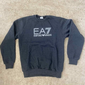 Mörkgrå EA7 Emporio Armani sweatshirt - Snygg mörkgrå sweatshirt från EA7 Emporio Armani med stort tryck på bröstet. Tröjan har rund hals, ribbade muddar och långärmad passform. Perfekt för dig som gillar stilrena märkesplagg med sportig vibe.