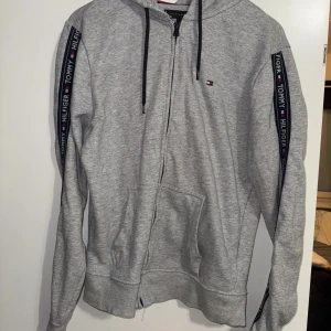 Grå hoodie Tommy Hilfiger M - Snygg grå hoodie från Tommy Hilfiger i storlek M. Tröjan har huva med svarta snören, dragkedja framtill och två fickor. På ärmarna finns ett band med Tommy Hilfiger-logga i marinblått, vitt och rött. Perfekt för chill eller streetwear.