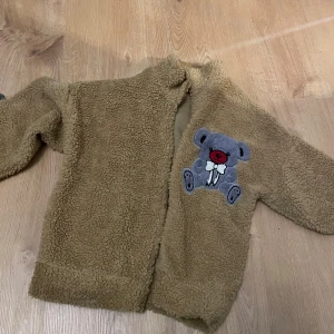 Ljus Khaki teddyjacka med nallepatch - Mysig ljuskhaki teddyjacka med fluffig yta och en stor broderad nallepatch på bröstet. Jackan har långärmade ärmar och är fodrad på insidan för extra komfort. Perfekt för dig som gillar gulliga detaljer och vill hålla dig varm.