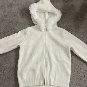 Vit stickad hoodie med fuskpäls Stockholmsstil - Mysig vit stickad hoodie med dragkedja och huva kantad med fluffig fuskpäls. Tröjan har ribbade muddar och nederkant, perfekt för kyliga dagar. Enkel och stilren design som passar dig som gillar en clean look. Den har två dragkedjor. 