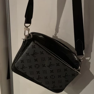 Svart axelväska - Snygg svart axelväska med klassiskt monogrammönster. Väskan har en rektangulär form, bred axelrem och silverfärgade dragkedjor. Tillverkad i skinn med diskret logga och blommor över hela ytan. Perfekt för dig som vill ha en stilren och lyxig look.