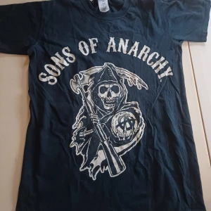 Sons of Anarchy svart t-shirt - Svart t-shirt med stort Sons of Anarchy-tryck på framsidan. Trycket visar en dödskalle med huva och gevär, samt texten 'Sons of Anarchy' i vitt. Klassisk rund hals och korta ärmar. Materialet är mjuk bomull, perfekt för dig som gillar serien och vill ha en cool look.