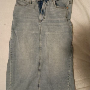 Ljusblå jeanskjol med klassisk look - Säljer en ljusblå jeanskjol med rak passform och klassiska fickor fram. Kjolen har en enkel design med synliga sömmar och dragkedja framtill. Perfekt för dig som gillar denim och vill ha en stilren kjol i garderoben. 2st fickor bak. Även kortare slits bak.