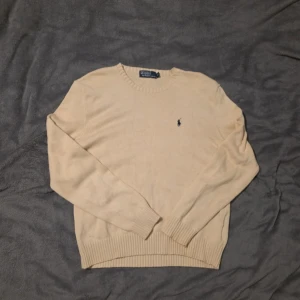 Beige stickad tröja Polo Ralph Lauren - Snygg beige stickad tröja från Polo Ralph Lauren med klassisk rund hals och broderad logga på bröstet. Tröjan har ribbade muddar vid ärmslut och nederkant, perfekt för dig som gillar stilrena och tidlösa plagg.