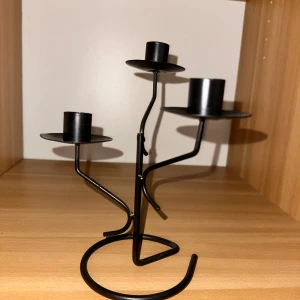 Kandelaber i svart metall ljushållare  - Bilderna visar en svart metall-ljusstake med plats för tre stearinljus. Modern och stilren design med böjda armar och rund fot. Passar utmärkt som dekorativ inredningsdetalj. Produkten ser ut att vara i gott skick utan synligt slitage eller defekter. Perfekt att dekorera hemmet med, göra det hemtrevligt och mysigt 🥰