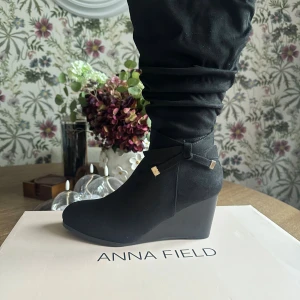 Svarta stövletter med kilklack från Anna Field - Oanvända svarta stövletter från Anna Field med kilklack och rund tå. Skorna har en dekorativ rosett med metalländar och dragkedja på sidan för enkel påtagning. Materialet är mocka-liknande syntet som ger en mjuk känsla och stilren look.