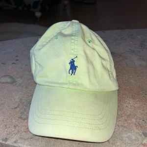 Ljusgul Polo Ralph Lauren keps -  ljusgul keps från Polo Ralph Lauren med klassisk blå broderad logga framtill och 'POLO' broderat baktill. Kepsen har böjd skärm och ventilerande hål, tillverkad i mjuk bomull. Perfekt för att lyfta din stil med en sportig touch.