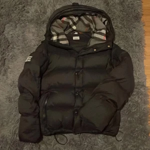 Svart Burberry pufferjacka med avtagbara ärmar - Svart pufferjacka från Burberry med avtagbara ärmar, så du kan bära den som väst. Jackan har huva med klassiskt rutigt Burberry-foder, dragkedja och knappar framtill samt två fickor. Stor Burberry London England-patch på ärmen.