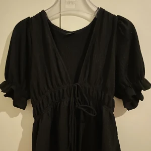 Svart kort klänning från Gina Tricot - Svart kort klänning från Gina Tricot med puffiga korta ärmar, v-ringning och knytning i midjan. Klänningen har en luftig känsla och lätt struktur i tyget, perfekt för en trendig och avslappnad look.