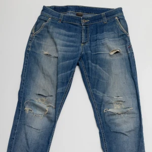 Blå slitna jeans från Dondup - Säljer ett par blå jeans från Dondup med snygga slitningar och trasiga detaljer på benen. Jeansen har klassisk femficksdesign, normal passform och är tillverkade i jeansmaterial. Bakfickorna har unika sömmar och läderpatch med logga. Storlek 14år passar dig som är runt 175cm lång.