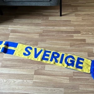 Sverige supporterhalsduk gul/blå - Supporterhalsduk i gult och blått med texten 'SVERIGE' på ena sidan och 'OFFICIAL SUPPORTER' på andra. Halsduken har svenska flaggan i ändarna och blå fransar. Perfekt för att visa ditt stöd på matchen.