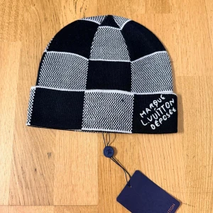 Mörkblåvit rutig mössa från Loui buitton - Snygg mörkblåvit mössa från Loui buitton med rutigt mönster och broderad text 'Marque L. Vuitton Deposee' på uppviket. Tillverkad i mjukt stickat material, perfekt för att lyfta din streetstyle. Mössan har märkets klassiska tagg och exklusiv känsla.